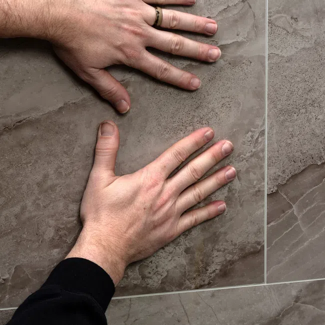 Two hands placed on large textured grey marble tiles showcasing surface and grout lines.