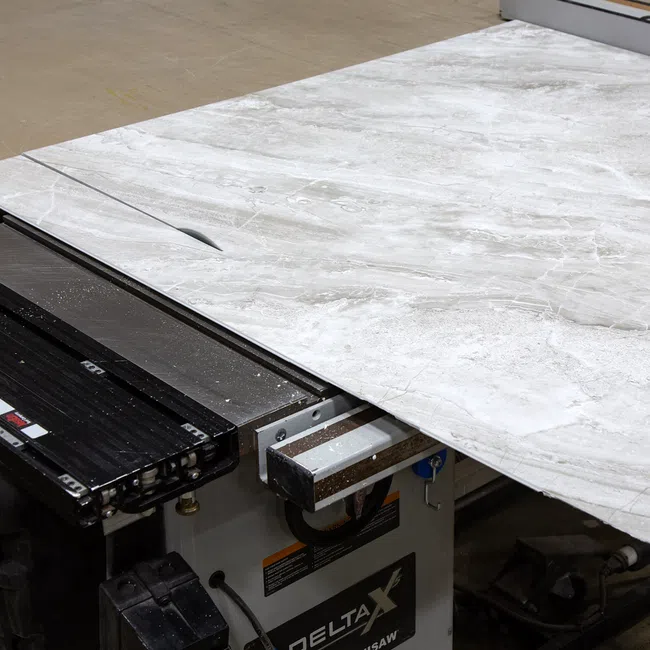 Large marble slab being cut on a Delta table saw in a workshop setting with dust and machinery visible.
