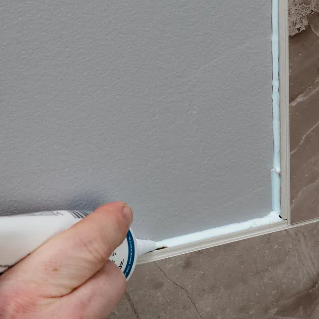 Hand applying white caulk to seal gap between wall and tiled floor for clean finish and moisture protection