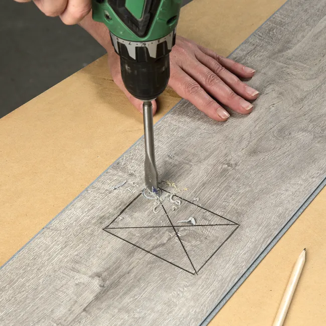 Person drilling a hole into a marked wooden plank using a green electric drill on a workbench.