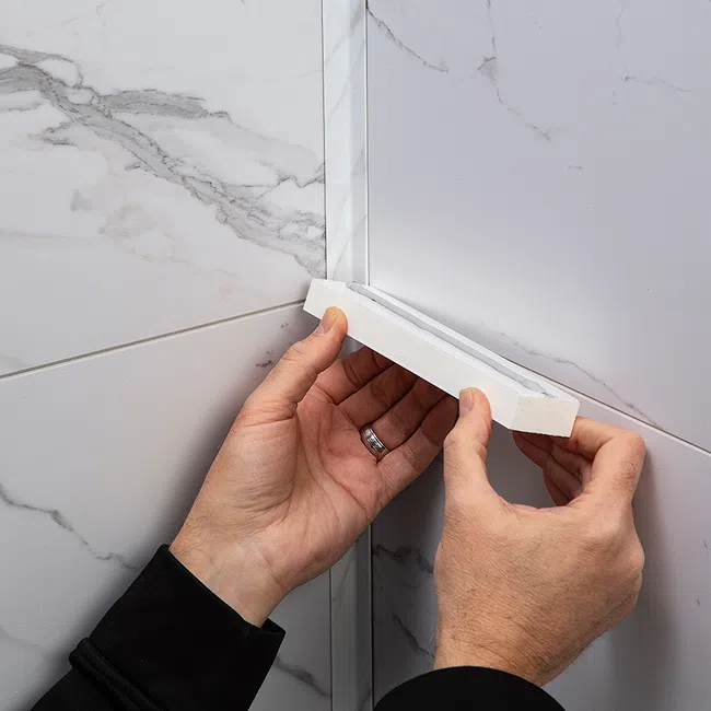 Hands installing white corner trim on marble-patterned wall tiles in a bathroom or kitchen setting