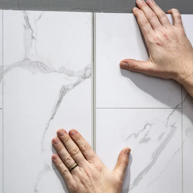 Hands installing large white marble-patterned ceramic floor tiles on a gray surface.