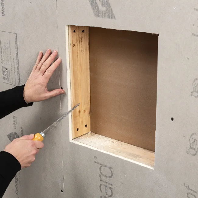 Person cutting drywall with a handsaw around a framed wall opening during home renovation.
