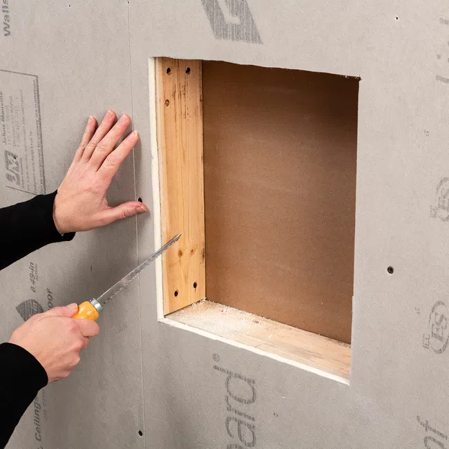 Person cutting drywall around a wooden window frame with a handsaw for installation or repair.