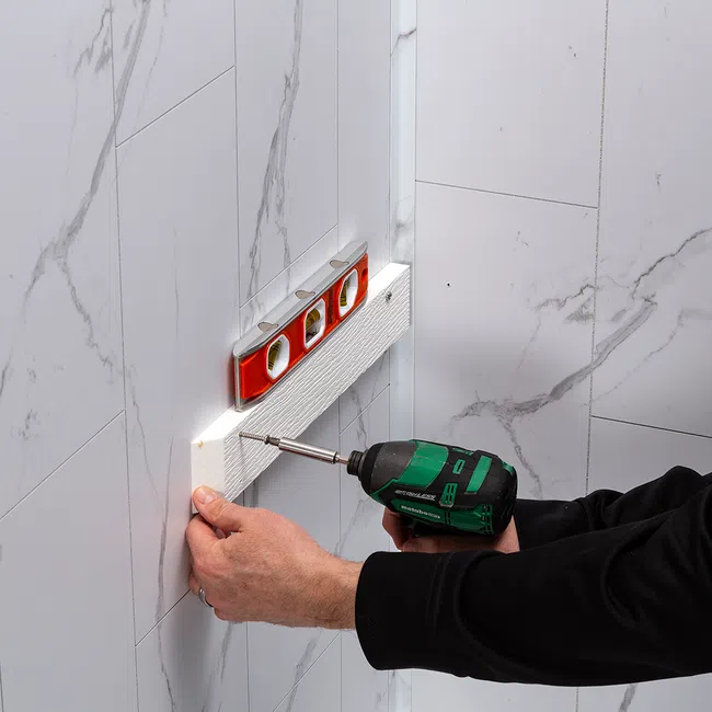 Person using a green cordless drill to screw a white textured panel on a wall with a red spirit level on top.