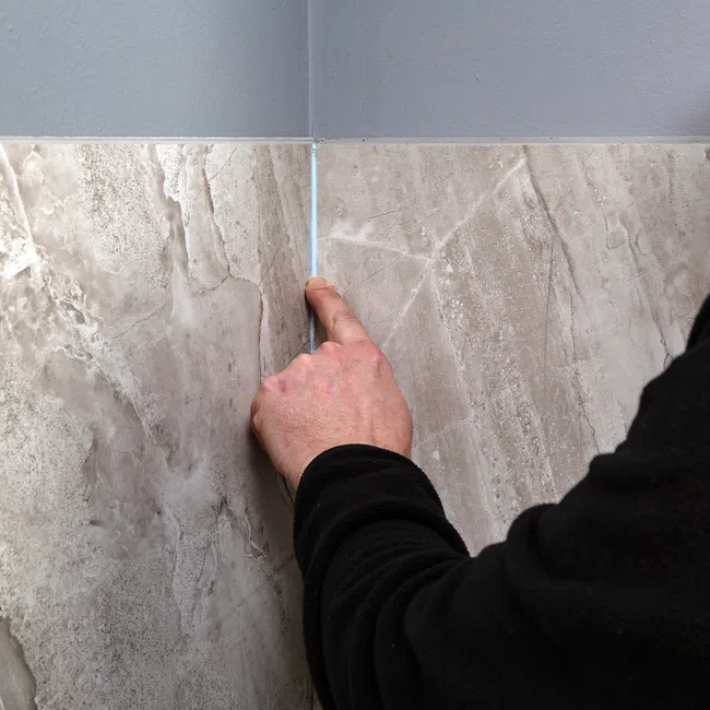 Person pointing at the thin gap between two beige marble wall tiles under a gray painted corner wall.