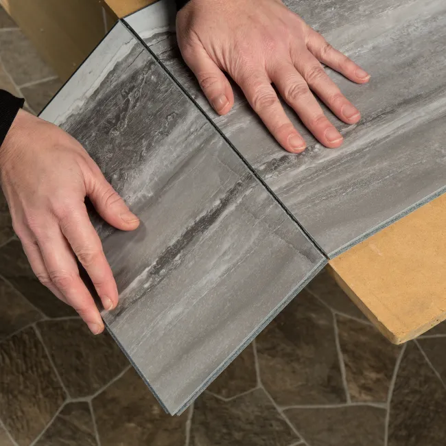 Hands installing gray textured vinyl flooring planks on a stone-patterned floor surface indoors.
