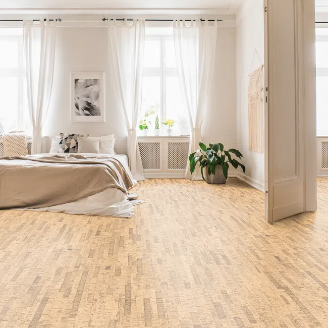 Bright bedroom with beige bedding, white curtains, wooden floor, green plant, and soft natural light.