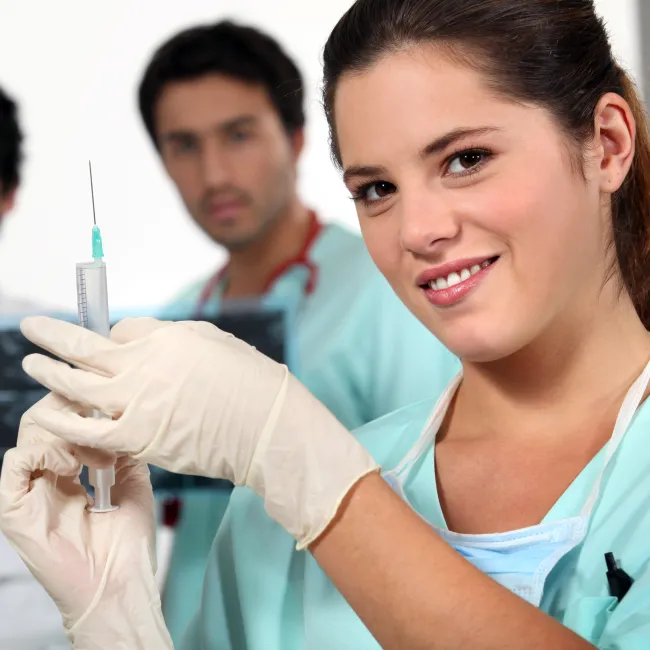 Smiling nurse wearing gloves prepares a syringe with two male doctors observing behind her.