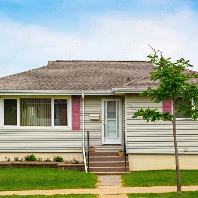 Single-story beige house with red shutters, chimney, front steps, small tree, and green lawn under blue sky.