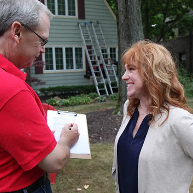 A man in a red shirt notes on a clipboard while talking to a woman outdoors in a residential area.