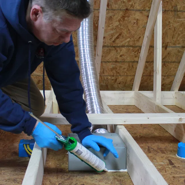 Worker in blue hoodie and gloves applying sealant to ventilation duct in attic framed with wood beams.
