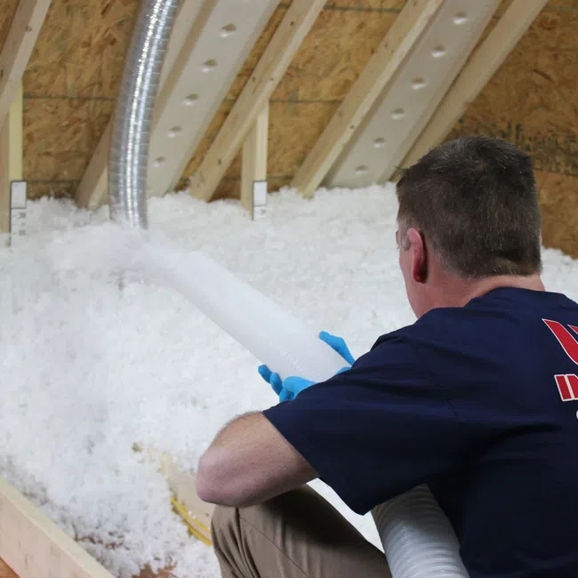 Worker installing loose-fill insulation in attic using hose to distribute material evenly.