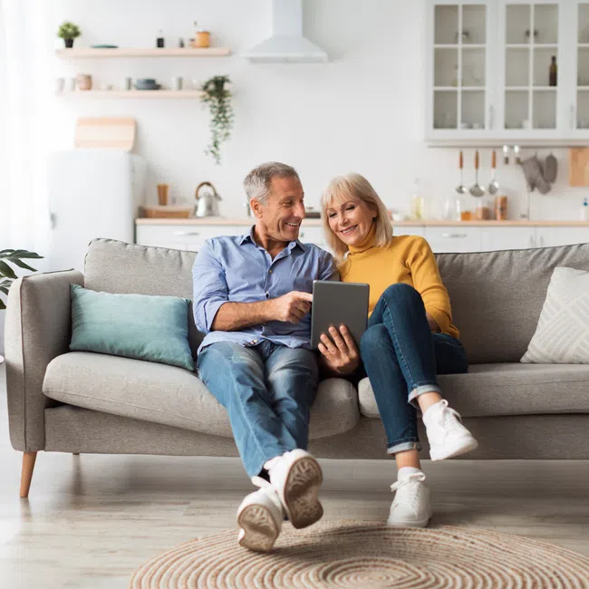 Smiling senior couple sitting on sofa using tablet in bright modern living room with cozy decor