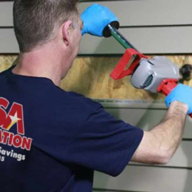 Worker wearing blue gloves using power tool to install beige siding on a building exterior.