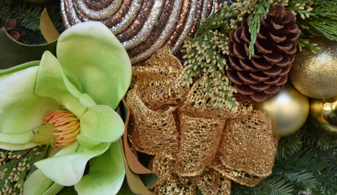 Close-up of green magnolia flower, pine cones, gold ribbon, and glittery ornaments on Christmas greenery.