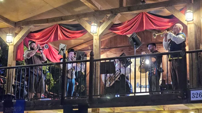 Five musicians playing brass instruments on a wooden balcony decorated with red curtains and lanterns at night.