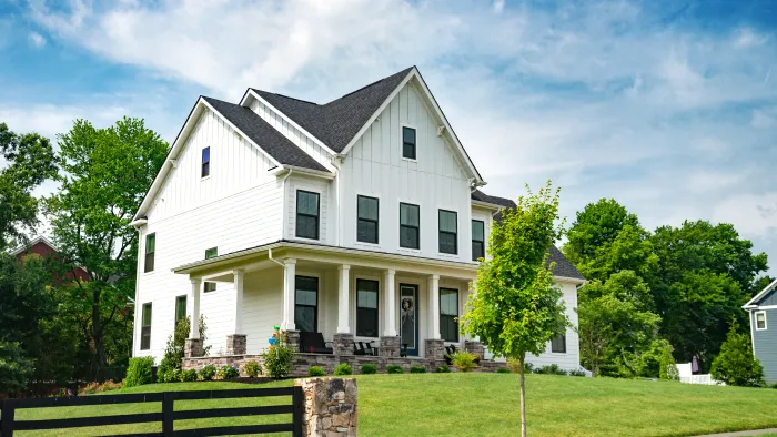 Large white two-story house with black windows and a spacious porch surrounded by green lawn and trees under a blue sky.