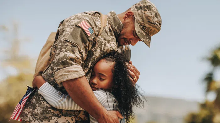 Soldier in uniform warmly hugging young girl holding American flag outdoors with blurred trees and sky background.