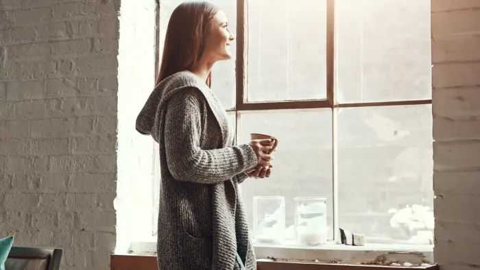 Woman in cozy sweater holding a cup, gazing out a large sunlit window in a comfortable living space.