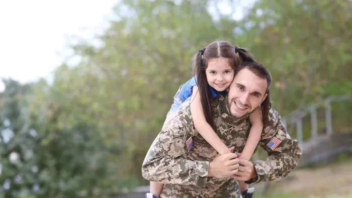 Smiling soldier in camouflage uniform carrying his young daughter on his back outdoors in a park.