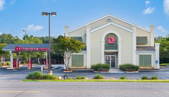 Red River Bank building with drive-thru lanes, surrounded by greenery under a partly cloudy sky.