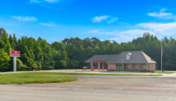 Small Red River Bank branch building with drive-thru lanes surrounded by trees and clear blue sky.