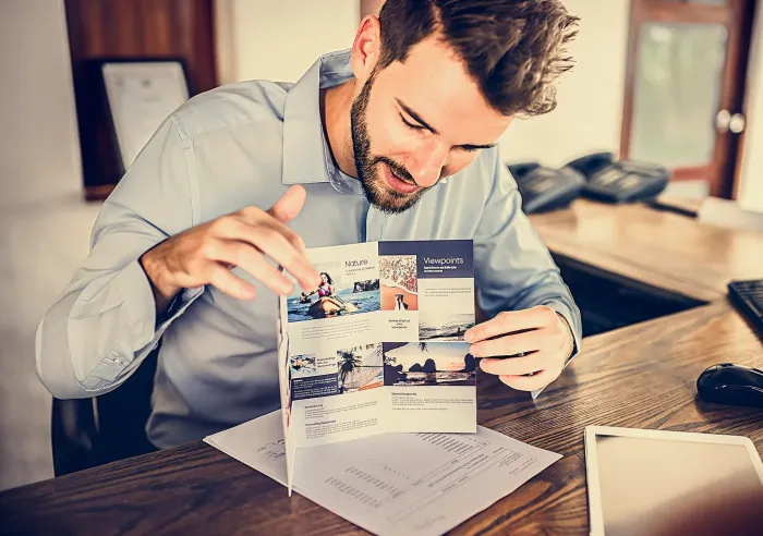 Man in office reading a travel brochure with images and text about nature and viewpoints at a wooden desk.