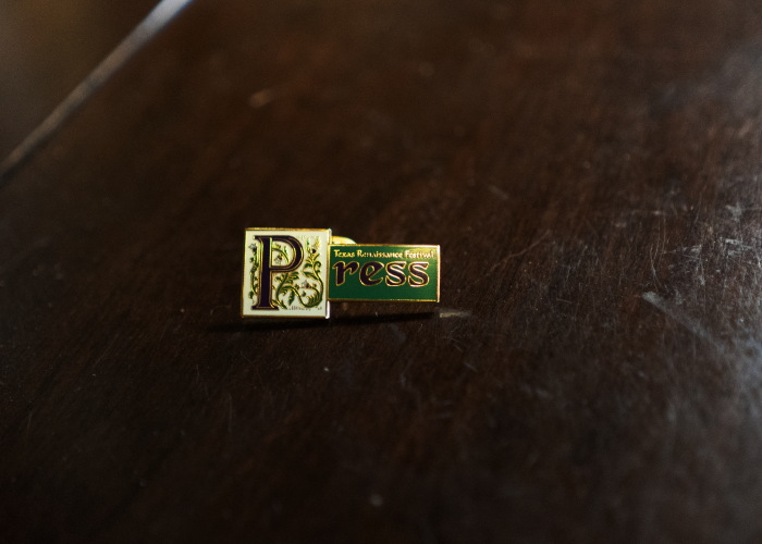 Enamel pin that reads PRESS in purple and gold lettering on a white and green background, sitting on a desk
