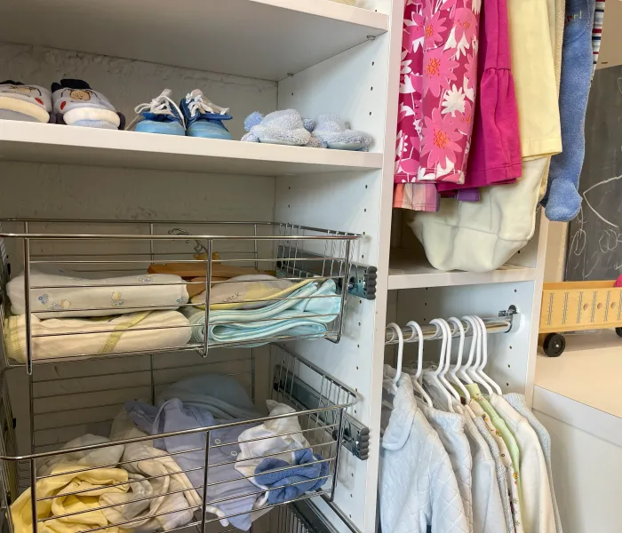 Organized baby closet with white shelves, folded baby clothes, baby shoes, hanging clothes and sliding metal bins with clothing