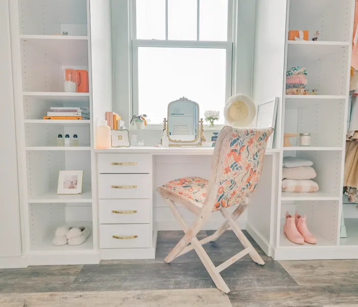 Walk in closet with white vanity area with floral cushioned chair, white shelves, glass doors, hanging clothes, slippers, sweaters and a window