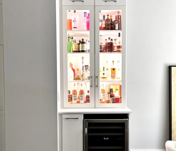 Illuminated custom wet bar with built-in white cabinets and glass doors with various liquor bottles and a mini fridge