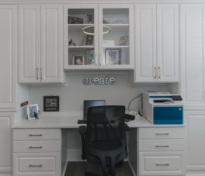 Modern home office with white cabinets, white desk, glass cabinet doors, laptop, printer, black executive chair and decorative create sign on wall.