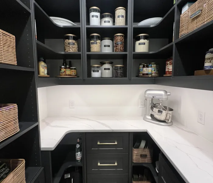 Kitchen pantry with white marble countertop, built-in dark brown shelves, drawers, baskets, jars and a silver Cuisinart mixer