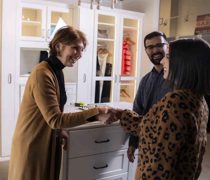 a female closet designer consulting with a man and woman who are standing in a walk in closet with white shelves, cabinets and glass cabinet doors