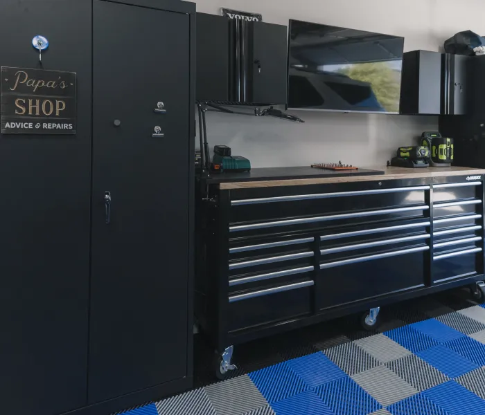 Organized garage workspace with black tool cabinets, workbench, TV, and blue and gray checkered flooring.