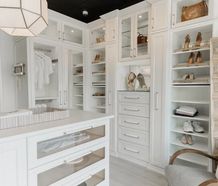 Custom designed walk-in closet with white island, white shelving, drawers, glass cabinets, accent lighting, hanging clothes, shoes and handbags