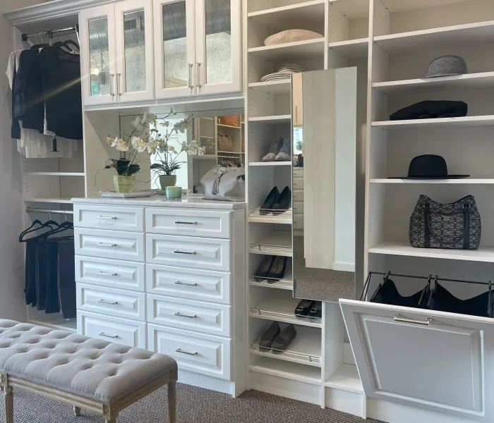 White walk-in closet with shelves, drawers, glass doors, a mirror, hamper and a tufted bench on carpeted floor