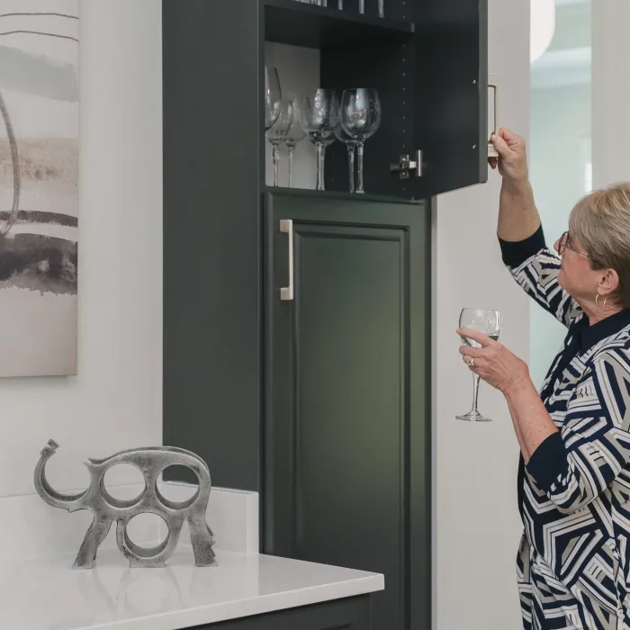 Woman wearing patterned dress holding a wine glass opening an upper kitchen cabinet with glassware inside.