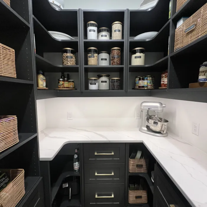 Organized kitchen pantry with white countertop, black cabinets and drawers, wicker baskets, glass jars and a silver Cuisinart mixer
