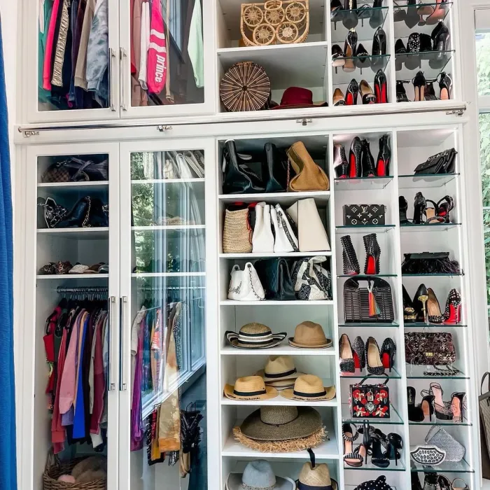Organized custom closet with glass doors displaying clothes, hats, handbags, and high-heeled shoes on white shelves.