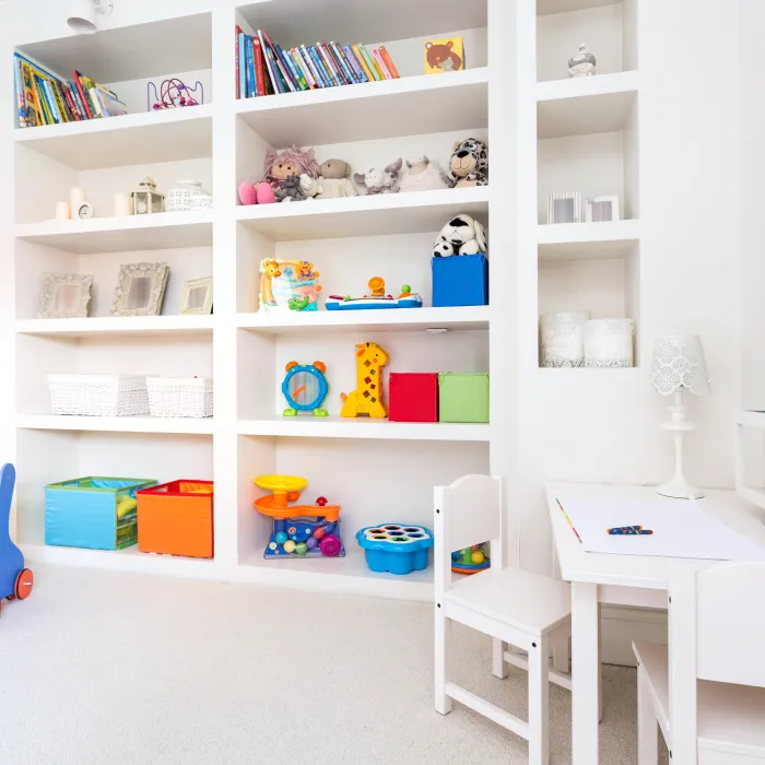 a childs playroom with built-in white shelves filled with books, stuffed toys and childrens toys and a small white desk with two white chairs and a blackboard on the wall