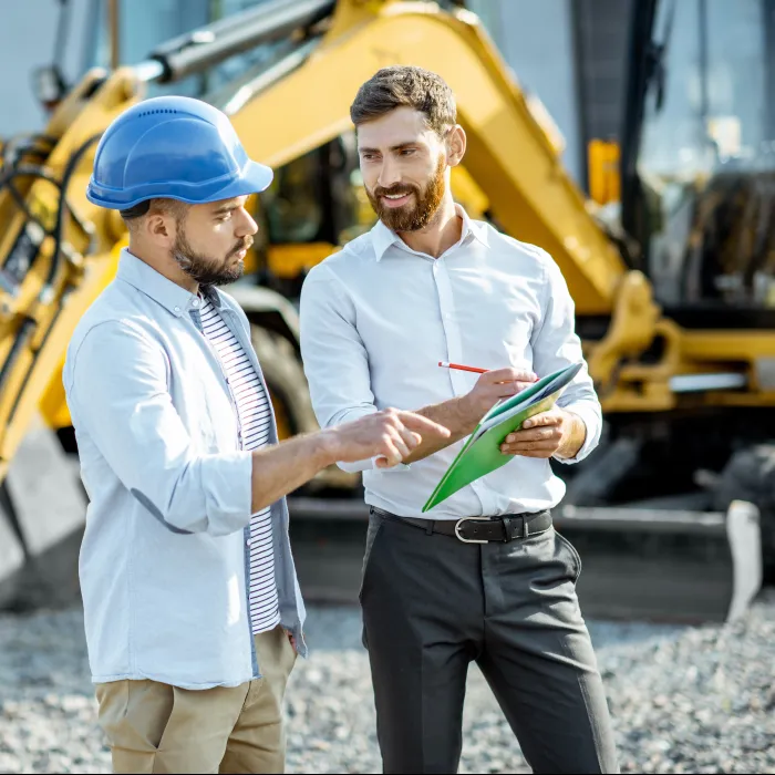Two men discuss construction plans at a worksite with heavy machinery in the background