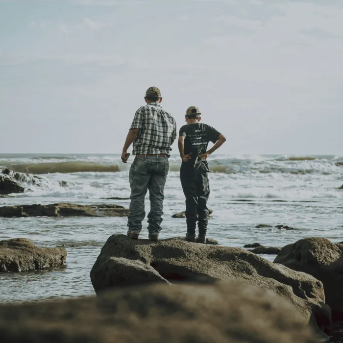 Two people stand on large rocks looking out at the ocean with waves and cloudy sky in the background.