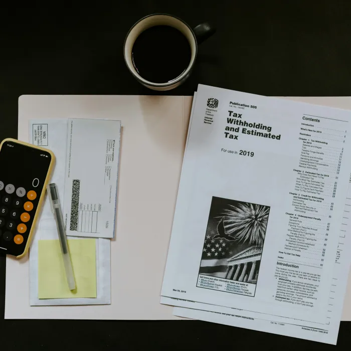 Desk with tax documents, calculator app on smartphone, envelopes, pen, sticky note, and a cup of coffee.