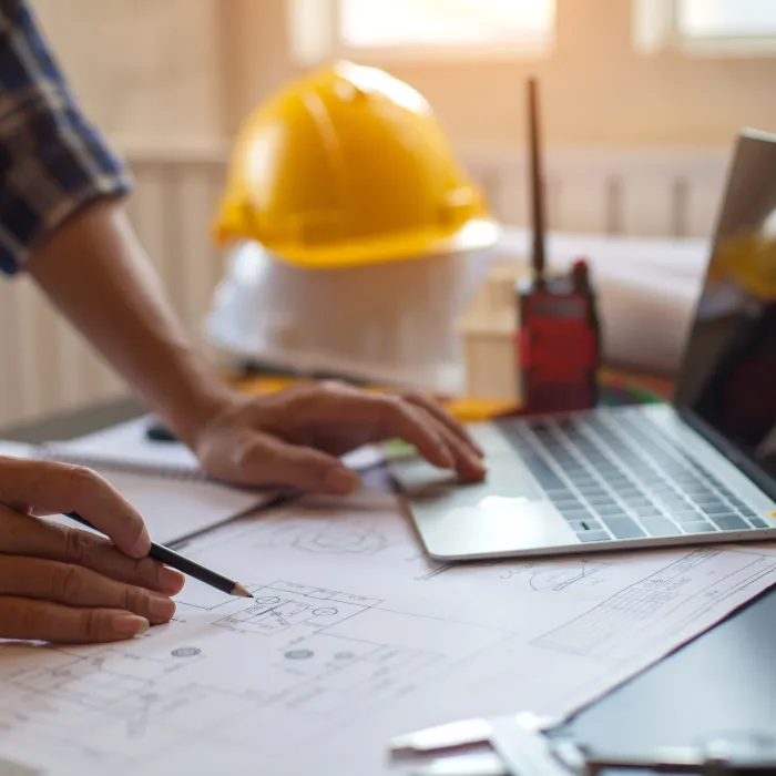 Engineer working on architectural blueprints with laptop, safety helmet, and walkie-talkie on desk in office