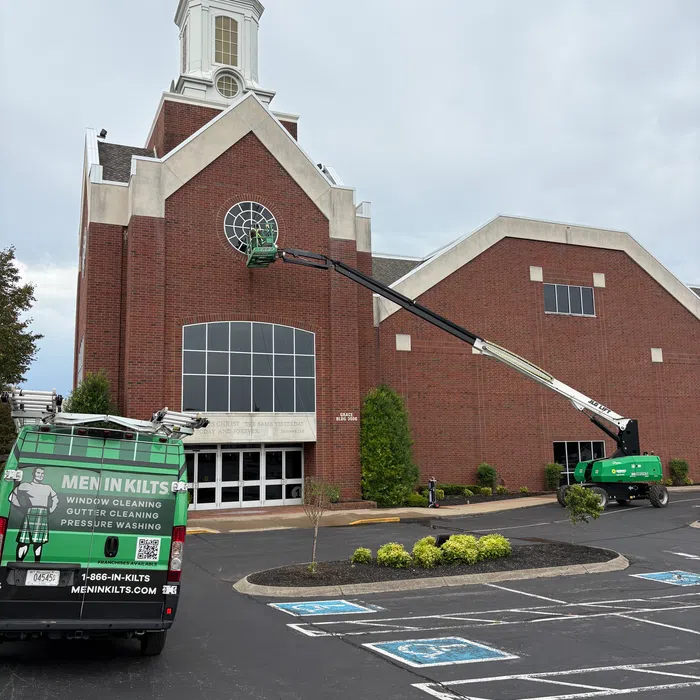 Green Men In Kilts van and lift cleaning stained glass window on red brick church on cloudy day.
