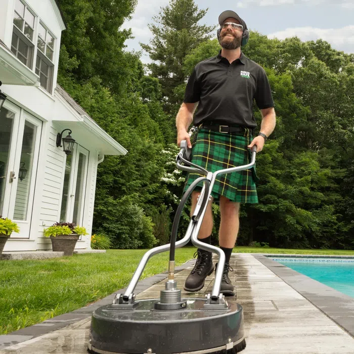 Man in kilt using pressure washer to clean poolside patio with house and trees in the background.