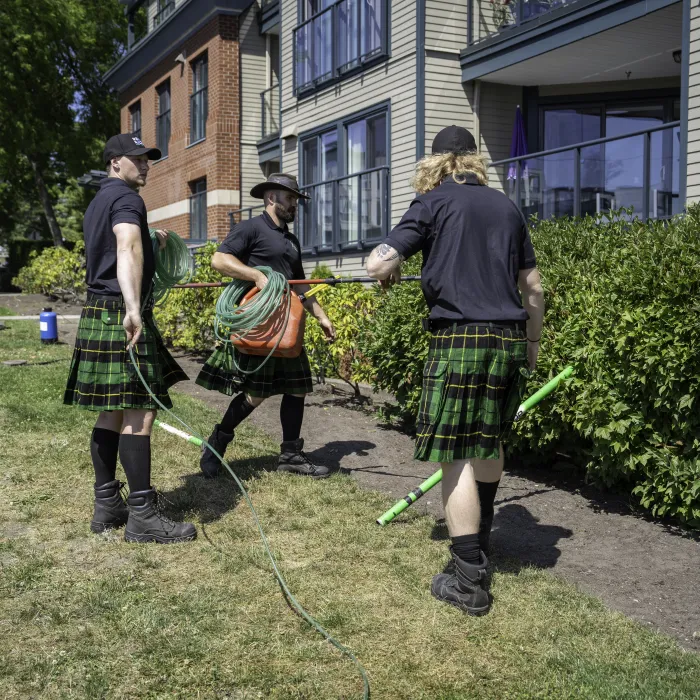 Three men in kilts preparing garden hoses outside residential buildings on a sunny day