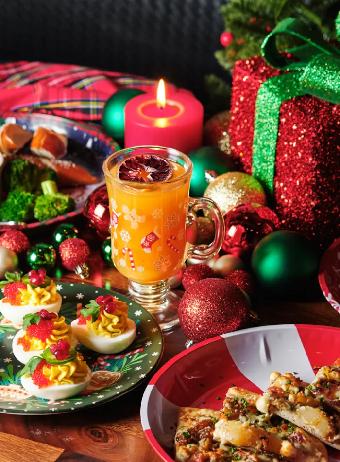 Festive holiday meal with deviled eggs, pizza slices, orange drink, Christmas decorations, and a sparkling gift box.